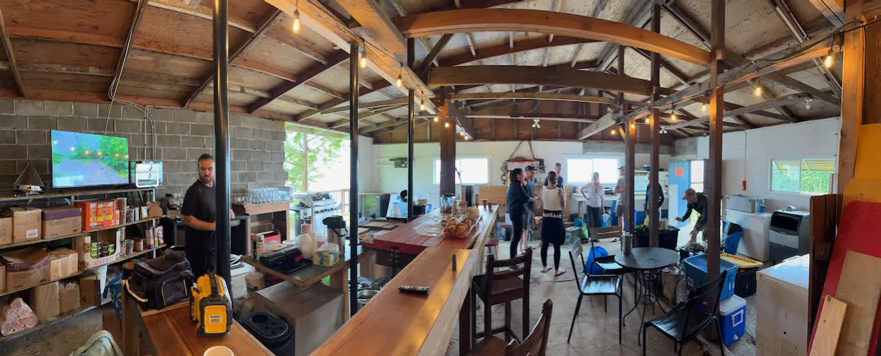 Panoramic interior view of the covered staging area at Woods End Landing with timber roof, string lights, long wooden bar, and paddlers gathering