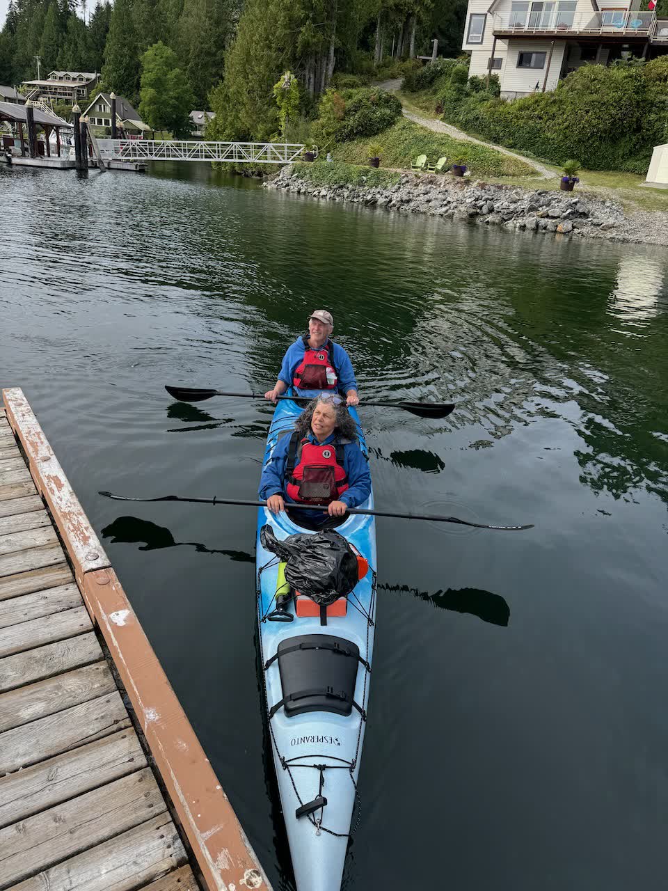 Two paddlers in a blue double sea kayak alongside a wooden dock on calm water in Bamfield