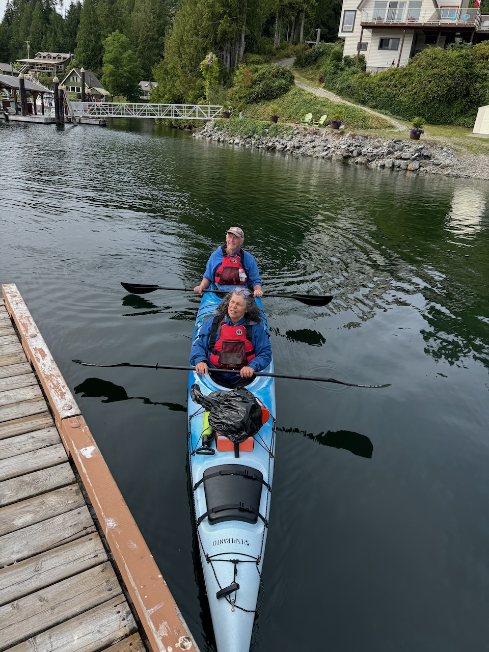 Two paddlers in a blue double sea kayak alongside a wooden dock on calm water in Bamfield
