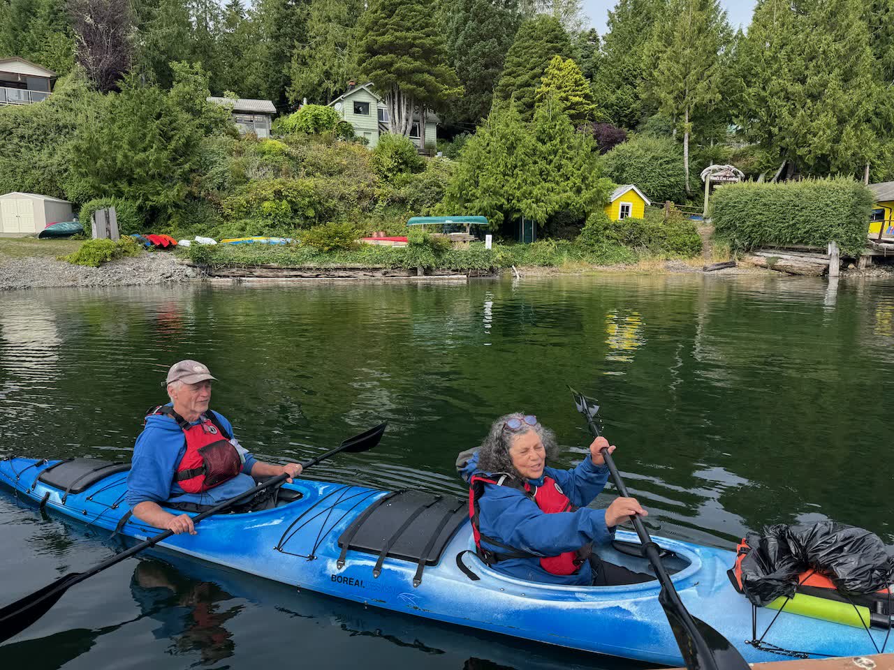 Two paddlers in a blue Boréal Design double sea kayak paddling on green water near a forested shoreline