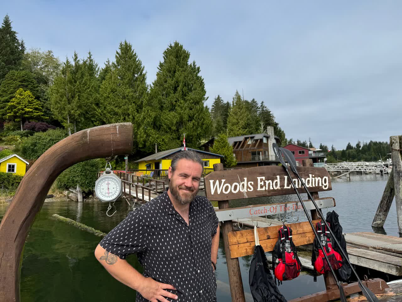 Person standing in front of the carved wooden Woods End Landing sign with the dock and yellow building behind